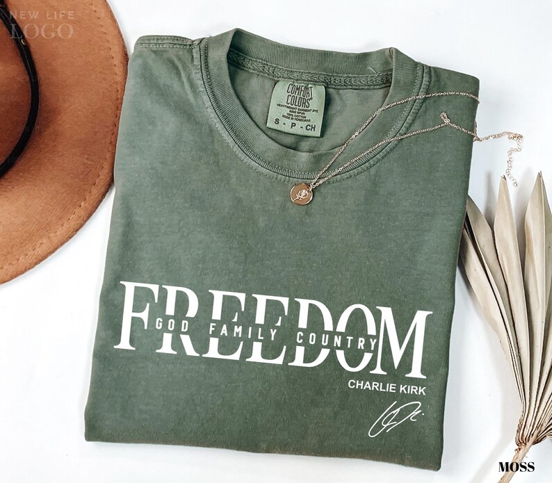 Puede incluir: Una camiseta verde musgo con la palabra "FREEDOM" impresa en blanco, junto con "GOD FAMILY COUNTRY" y la firma "CHARLIE KIRK". Un collar dorado con un colgante descansa sobre la camiseta. Un sombrero marr&oacute;n y una planta seca est&aacute;n en el fondo.