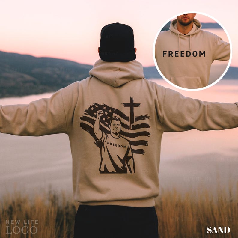 May include: Sand-colored hoodie with the word "FREEDOM" printed on the front and back. The back features a graphic of a person raising a fist in front of an American flag and a cross. The hoodie is a neutral color.