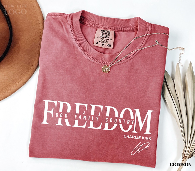 Puede incluir: Camiseta de color carmes&iacute; con la palabra "FREEDOM" impresa en blanco, junto con "GOD FAMILY COUNTRY" y "CHARLIE KIRK". Un collar dorado con un colgante descansa sobre la camiseta. La camiseta tiene una etiqueta Comfort Colors.