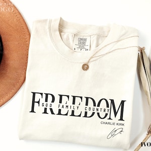 Puede incluir: Una camiseta de color marfil con la palabra "FREEDOM" impresa en letras negras grandes y en negrita. Debajo de la palabra "FREEDOM" est&aacute;n las palabras "GOD FAMILY COUNTRY" y el nombre "CHARLIE KIRK" en letras negras m&aacute;s peque&ntilde;as. Un collar de oro descansa sobre la camisa.