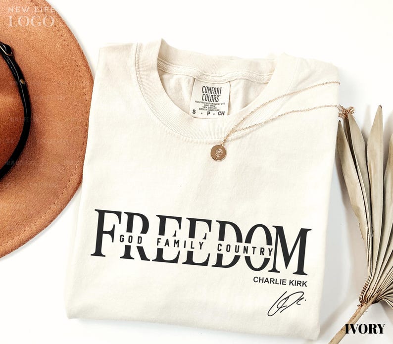 Puede incluir: Una camiseta de color marfil con la palabra "FREEDOM" impresa en letras negras grandes y en negrita. Debajo de la palabra "FREEDOM" est&aacute;n las palabras "GOD FAMILY COUNTRY" y el nombre "CHARLIE KIRK" en letras negras m&aacute;s peque&ntilde;as. Un collar de oro descansa sobre la camisa.