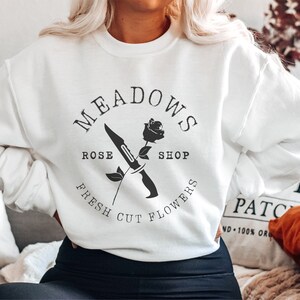 May include: White sweatshirt with a black graphic design that says "Meadows Rose Shop Fresh Cut Flowers" with a rose and a knife.