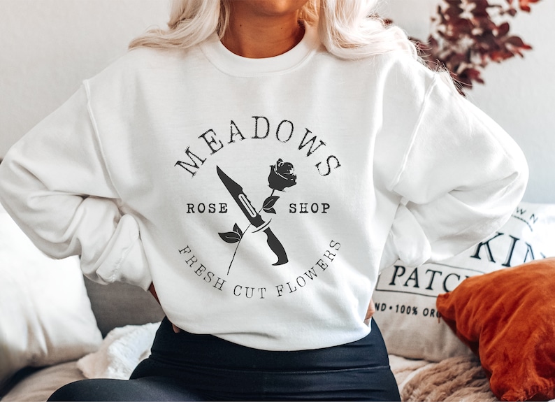 May include: White sweatshirt with a black graphic design that says "Meadows Rose Shop Fresh Cut Flowers" with a rose and a knife.