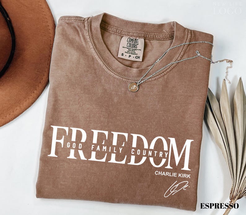 Puede incluir: Camiseta color espresso con la palabra "FREEDOM" en letras blancas. Debajo de la palabra "FREEDOM" est&aacute;n las palabras "GOD FAMILY COUNTRY" y el nombre "CHARLIE KIRK" con una firma. Un collar plateado descansa sobre la camiseta.
