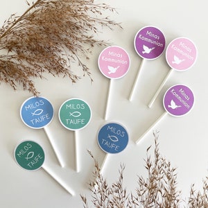 Könnte beinhalten: Sechs runde Tortendekorationen mit verschiedenen Designs. Vier Dekorationen sind blau und grün mit einem Fischsymbol und dem Text "MILOS TAUFE". Zwei Dekorationen sind rosa und lila mit einem Taubensymbol und dem Text "Minas Kommunion".