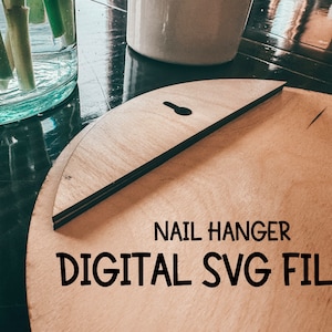 May include: A light brown wooden circle with a nail hanger cut out. The circle has a hole at the top for hanging. The text "NAIL HANGER DIGITAL SVG FILE!" is printed on the circle.