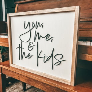 May include: A white wooden sign with black cursive lettering that reads "You, Me, & The Kids". The sign is leaning against a wooden piano.