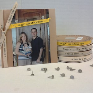 May include: A brown scrapbook with a photo of a family of three. The scrapbook has a yellow ribbon with the text "a dream come true" on it. The photo shows a woman, a man, and a baby. The text "no longer under construction" is on the scrapbook. There are several spools of white ribbon with text on them.