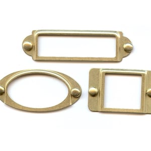 May include: Three gold metal label holders with rounded edges and small decorative knobs. The label holders are in different shapes: rectangular, oval, and square.