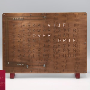 Op de afbeelding: Een houten woordklok met een koperen afwerking. De klok geeft de tijd weer met Nederlandse woorden voor de uren en minuten. De klok staat op een standaard met rode poten. De klok is gelabeld "WordClock by StarkAllister".