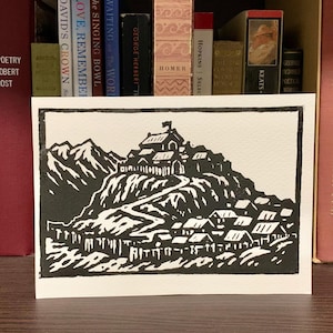 May include: Black and white linocut print of a mountain village with a flag atop the highest building. The print is displayed on a shelf in front of a collection of books with various spines and colors.