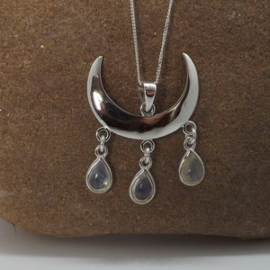 May include: A silver-toned crescent moon pendant necklace with three teardrop-shaped gemstones. The necklace hangs on a delicate chain. The gemstones have a subtle, iridescent quality. The pendant is displayed on a brown, textured surface.