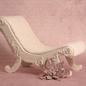 May include: A white, ornately carved chaise lounge with a pink, plush cushion. A pink flower with silver accents sits in front of the chaise lounge.