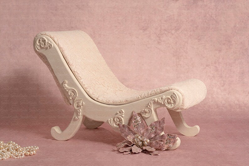 May include: A white, ornately carved chaise lounge with a pink, plush cushion. A pink flower with silver accents sits in front of the chaise lounge.