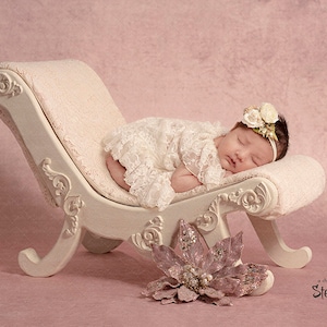May include: A sleeping baby girl wearing a white lace dress and a flower headband lies on a white, ornate chaise lounge. A pink sparkly flower is in the foreground.