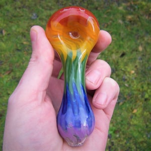 May include: A rainbow glass pipe with a rounded bowl and a long, narrow stem. The pipe is made of clear glass with streaks of red, orange, yellow, green, blue, and purple.