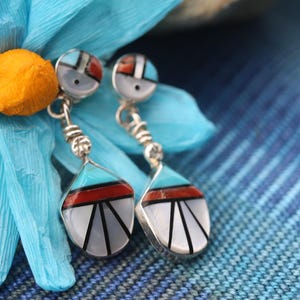May include: A pair of dangle earrings with turquoise, red, white, and black inlay. The earrings are made of sterling silver and have a geometric design.