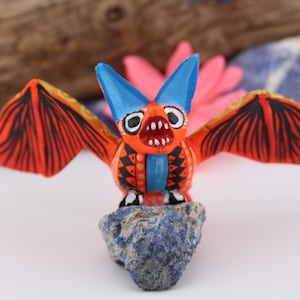 May include: A hand-painted, orange bat figurine with blue ears and wings, perched on a blue stone. The bat has black wing details and a wide open mouth. The background includes a wooden log and pink flower.