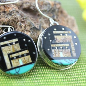 May include: A pair of round silver earrings with black, brown, and turquoise inlay. The earrings feature a geometric design.