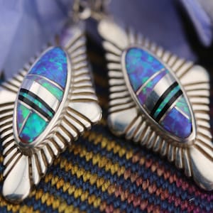 Könnte beinhalten: Ein Paar silberne Ohrringe mit einem geometrischen Design, in dessen Mitte ein blauer und grüner Opal-Stein sitzt. Die Ohrringe haben eine Diamantform und sind mit filigranen Details verziert.