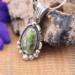 May include: A silver pendant necklace with a teardrop-shaped, green and white stone. The pendant is set in a decorative silver frame with beaded and rope-like details. The necklace is displayed on a textured surface.