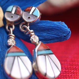 May include: Close-up of silver earrings with inlaid gemstones. The earrings feature a teardrop design with red, turquoise, and white accents. The earrings are displayed on a blue fabric with a red background.