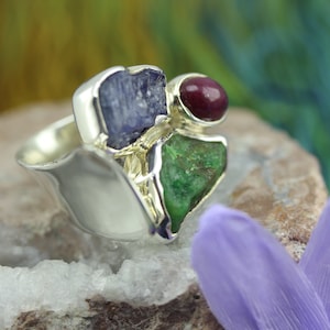 May include: A silver ring with three gemstones: a blue sapphire, a green emerald, and a red ruby. The ring is set on a rock with a purple flower.