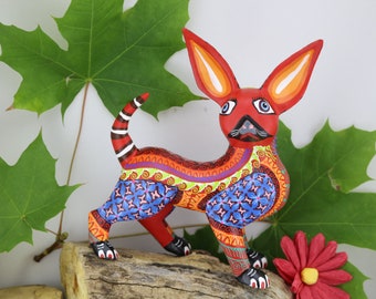 May include: A hand-painted wooden figurine of a red chihuahua with blue, green, orange, and white details. The figurine is standing on a wooden log with green leaves in the background.