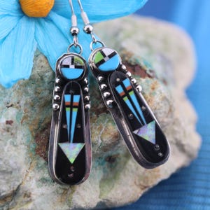 May include: A pair of dangle earrings with a geometric design. The earrings are made of silver and feature turquoise, black, and white stones.