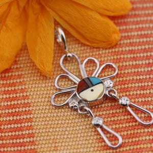 May include: A silver pendant with a flower design, featuring a central circular inlay with turquoise, red, and cream colors. The pendant has two dangling teardrop-shaped accents with small clear stones. An orange flower rests in the background.