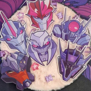 Pode incluir: Coleção de chaveiros de personagens de desenhos animados em vários tons de roxo, rosa e cinza. Cada chaveiro apresenta um design de rosto de robô diferente com argolas prateadas. Os chaveiros estão dispostos sobre uma superfície fofa e clara.