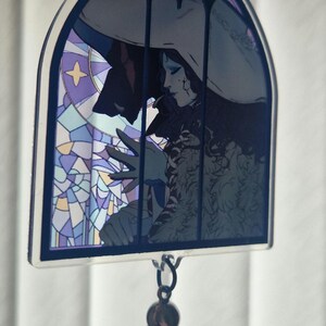 K&ouml;nnte beinhalten: Ein Anh&auml;nger im Glasmalerei-Stil mit einer gotischen Illustration einer Figur mit Hut, mit einer dunklen Silhouette und einer blauen Quaste. Der Anh&auml;nger hat einen dunkelblauen Rand und eine Kette zum Aufh&auml;ngen.