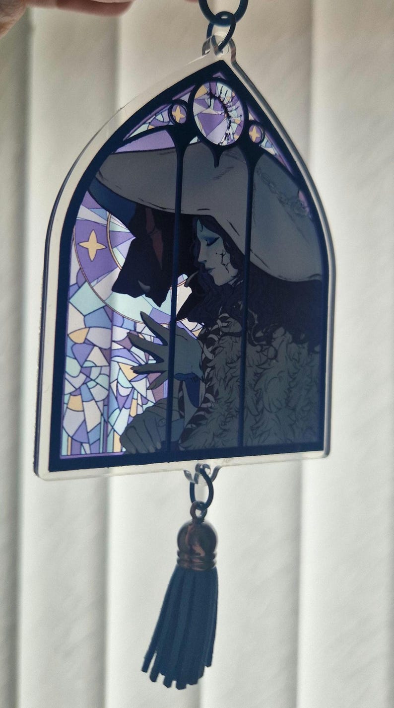 K&ouml;nnte beinhalten: Ein Anh&auml;nger im Glasmalerei-Stil mit einer gotischen Illustration einer Figur mit Hut, mit einer dunklen Silhouette und einer blauen Quaste. Der Anh&auml;nger hat einen dunkelblauen Rand und eine Kette zum Aufh&auml;ngen.