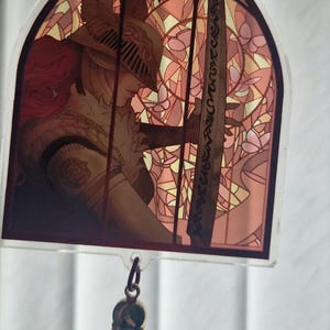 K&ouml;nnte beinhalten: Ein Acryl-Charm im Buntglas-Stil, das eine Kriegerfigur mit einem Schwert vor einem rosa-gelben Blumenmuster zeigt. Es hat eine dunkelrote Quaste und einen Metallverschluss.