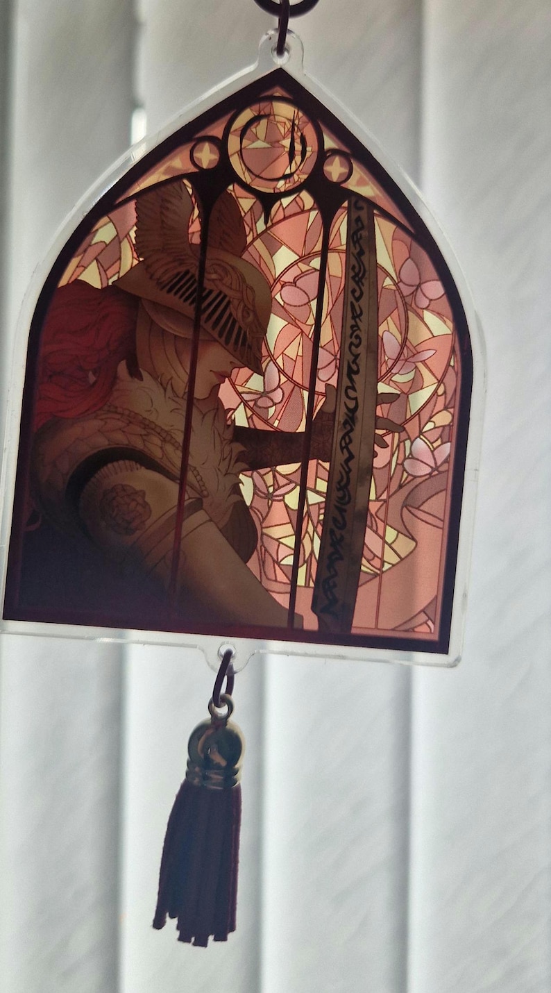 K&ouml;nnte beinhalten: Ein Acryl-Charm im Buntglas-Stil, das eine Kriegerfigur mit einem Schwert vor einem rosa-gelben Blumenmuster zeigt. Es hat eine dunkelrote Quaste und einen Metallverschluss.