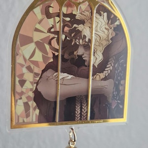 K&ouml;nnte beinhalten: Ein klarer Acryl-Anh&auml;nger mit goldenem Rand und einem Buntglasdesign mit einer Figur mit lockigem Haar. Der Anh&auml;nger hat eine goldene Quaste. Das Kunstwerk ist in Braun-, Beige- und Goldt&ouml;nen gehalten.