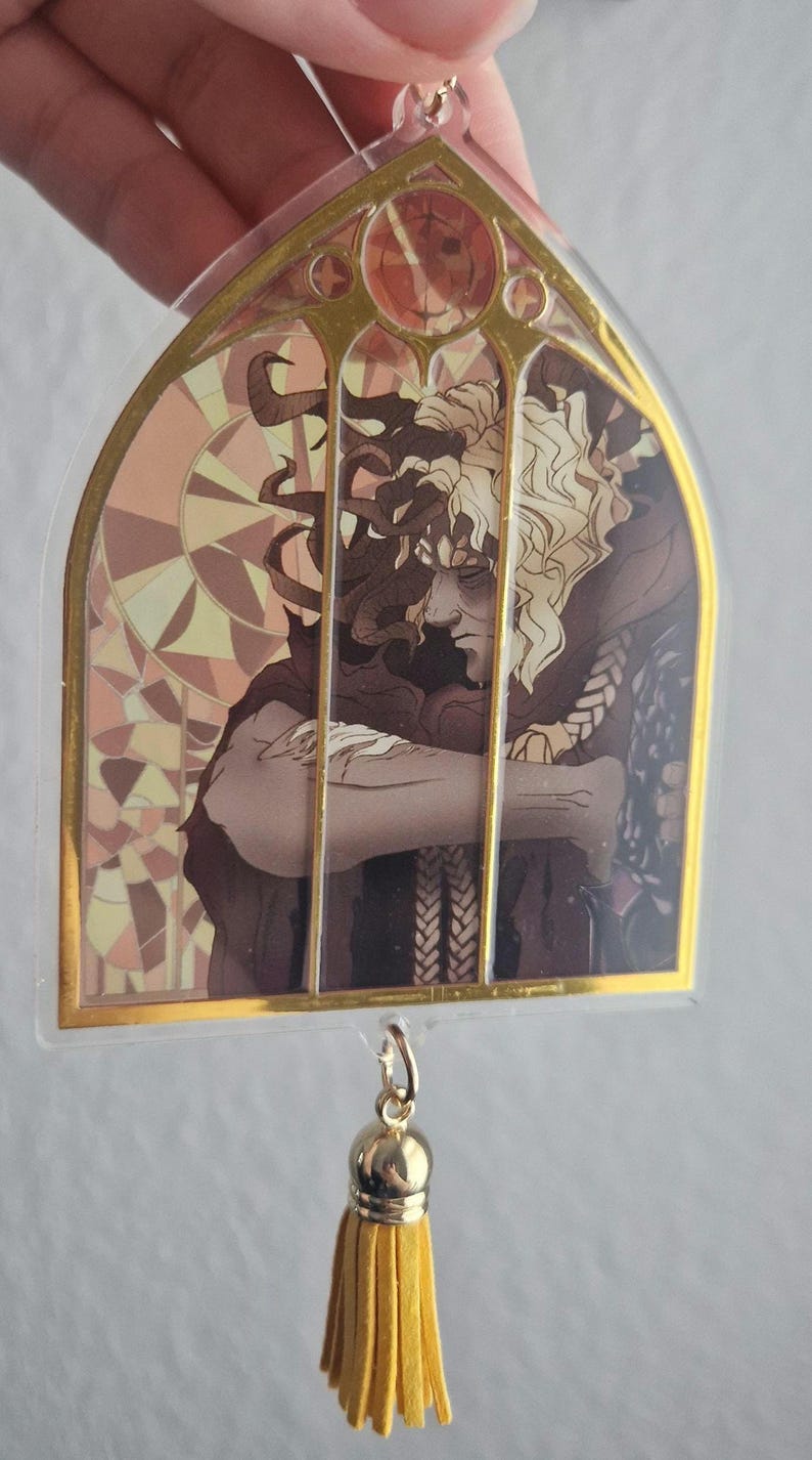 K&ouml;nnte beinhalten: Ein klarer Acryl-Anh&auml;nger mit goldenem Rand und einem Buntglasdesign mit einer Figur mit lockigem Haar. Der Anh&auml;nger hat eine goldene Quaste. Das Kunstwerk ist in Braun-, Beige- und Goldt&ouml;nen gehalten.