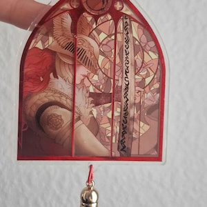 K&ouml;nnte beinhalten: Rot gerahmter Acryl-Anh&auml;nger mit einer Buntglas-Illustration eines Charakters mit Helm und rotem Haar. Der Anh&auml;nger hat eine rote Quaste und eine goldfarbene Kappe. Das Design ist detailliert mit warmen Farben.
