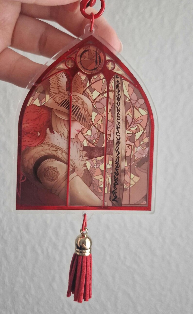 K&ouml;nnte beinhalten: Rot gerahmter Acryl-Anh&auml;nger mit einer Buntglas-Illustration eines Charakters mit Helm und rotem Haar. Der Anh&auml;nger hat eine rote Quaste und eine goldfarbene Kappe. Das Design ist detailliert mit warmen Farben.