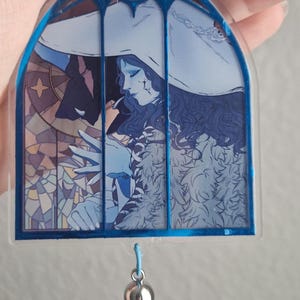 K&ouml;nnte beinhalten: Ein blau-wei&szlig;er Acryl-Schl&uuml;sselanh&auml;nger mit einem Buntglasfenster-Design. Das Kunstwerk zeigt eine Figur mit wei&szlig;em Hut und blauer Kleidung. Am unteren Ende h&auml;ngt eine blaue Quaste.