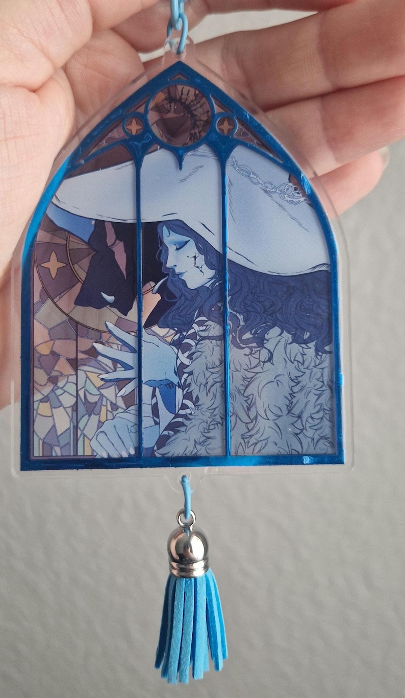 K&ouml;nnte beinhalten: Ein blau-wei&szlig;er Acryl-Schl&uuml;sselanh&auml;nger mit einem Buntglasfenster-Design. Das Kunstwerk zeigt eine Figur mit wei&szlig;em Hut und blauer Kleidung. Am unteren Ende h&auml;ngt eine blaue Quaste.