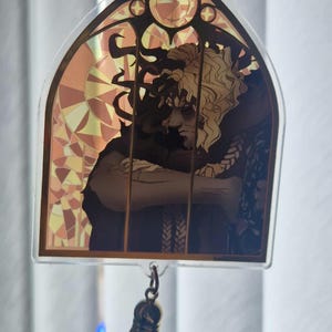 K&ouml;nnte beinhalten: Ein klarer Acryl-Schl&uuml;sselanh&auml;nger mit einem Buntglas-Design, das eine Charakterillustration zeigt. Das Kunstwerk zeigt eine Person mit lockigem blondem Haar und dunkler Kleidung. Eine braune Quaste h&auml;ngt von unten. Der Schl&uuml;sselanh&auml;nger wird von einer Hand gehalten.