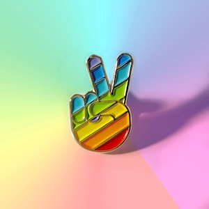 May include: A gold-colored metal pin in the shape of a hand making the peace sign. The hand is filled with a rainbow of colors, including red, orange, yellow, green, blue, and purple. The pin has a glossy finish.