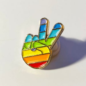 May include: A colorful enamel pin in the shape of a hand making the peace sign. The pin features a rainbow design with red, orange, yellow, green, blue, and purple stripes, outlined in gold. The pin is a symbol of peace and love.