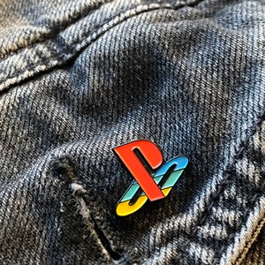 以下が含まれることがあります： 濃いグレーのデニムの背景に、赤、青、黄色のプレイステーションロゴのピン
