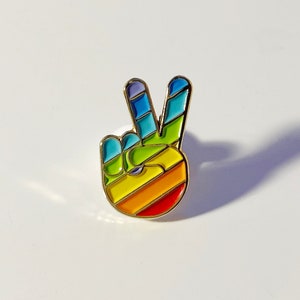 May include: A colorful enamel pin in the shape of a hand making the peace sign. The pin features a rainbow design with red, orange, yellow, green, blue, and purple stripes. The pin has a gold-colored border.