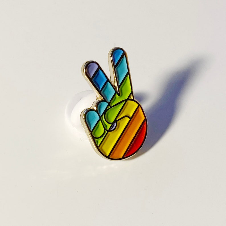 May include: A colorful peace sign pin with a gold border. The pin features a hand gesture with the fingers forming a V shape, filled with a rainbow of colors including red, orange, yellow, green, blue, and purple. The pin is set against a white background.
