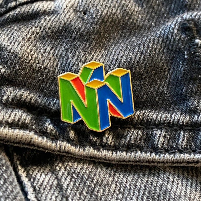 Peut inclure: Une &eacute;pingle en &eacute;mail color&eacute;e repr&eacute;sentant le logo Nintendo 64. L'&eacute;pingle a une bordure dor&eacute;e avec des sections vertes, rouges et bleues. L'&eacute;pingle est pos&eacute;e sur un fond en denim textur&eacute;.