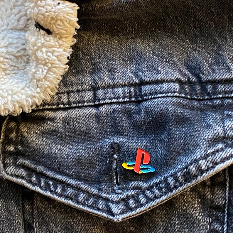 以下が含まれることがあります： ポケットにプレイステーションのロゴピンが付いた、茶色のふわふわした襟のある黒いデニムジャケットです。