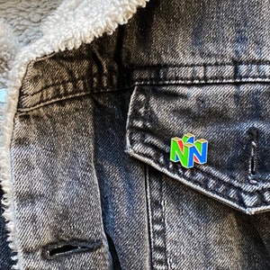 Peut inclure: &Eacute;pinglette color&eacute;e en &eacute;mail repr&eacute;sentant le logo Nintendo 64 vert, rouge et bleu. L'&eacute;pinglette est fix&eacute;e sur une veste en jean noire avec une doublure blanche et duveteuse.